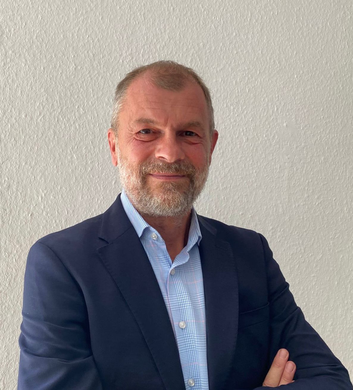 Ein älterer Mann mit grauem Bart lächelt freundlich im blauen Sakko vor hellem Hintergrund